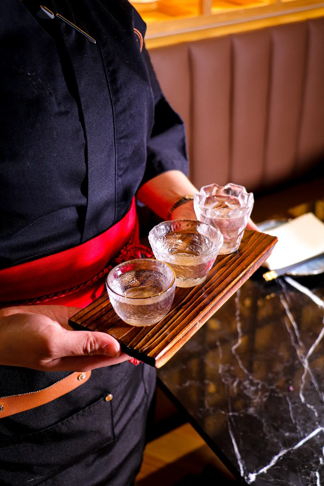 A server in a red sash carrying a flight of sake on a wooden board