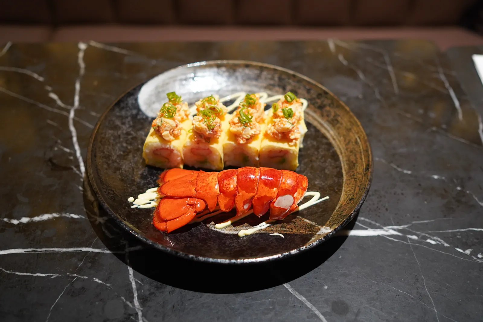 The Lobster & Crab Roll in egg crêpe beside a lobster tail, on a black plate